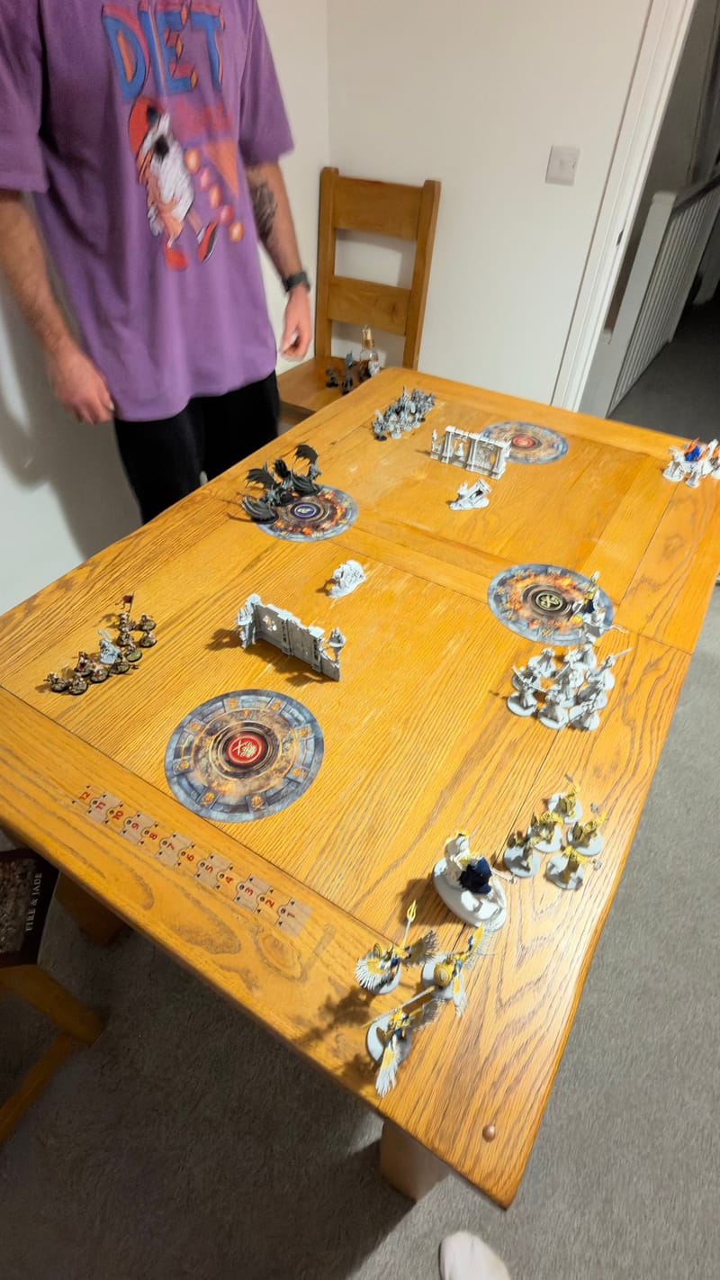 The Border War table ready for the game, with Peter standing at the far end: four round objective markers, ruined walls and both armies set up on a wooden dining table.