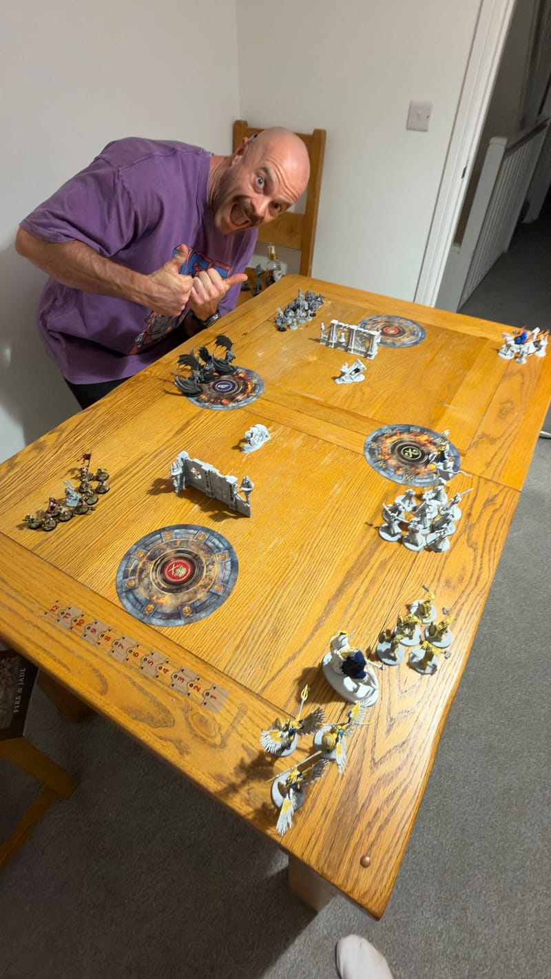Peter grinning and giving a big double thumbs up at the end of a wooden table set up for the game, with round objective markers, ruined walls and both armies in place.