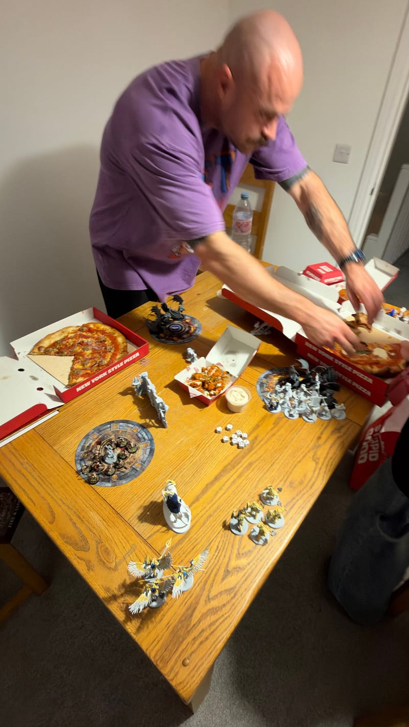 Peter leaning across the battle table to take a slice from an open pizza box, with more pizza and a side dish among the models and objective markers.