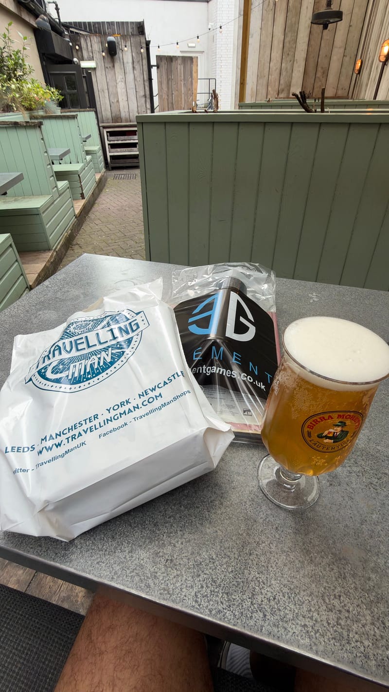 A Travelling Man bag and an Element Games bag on a metal pub table, next to a pint of lager.