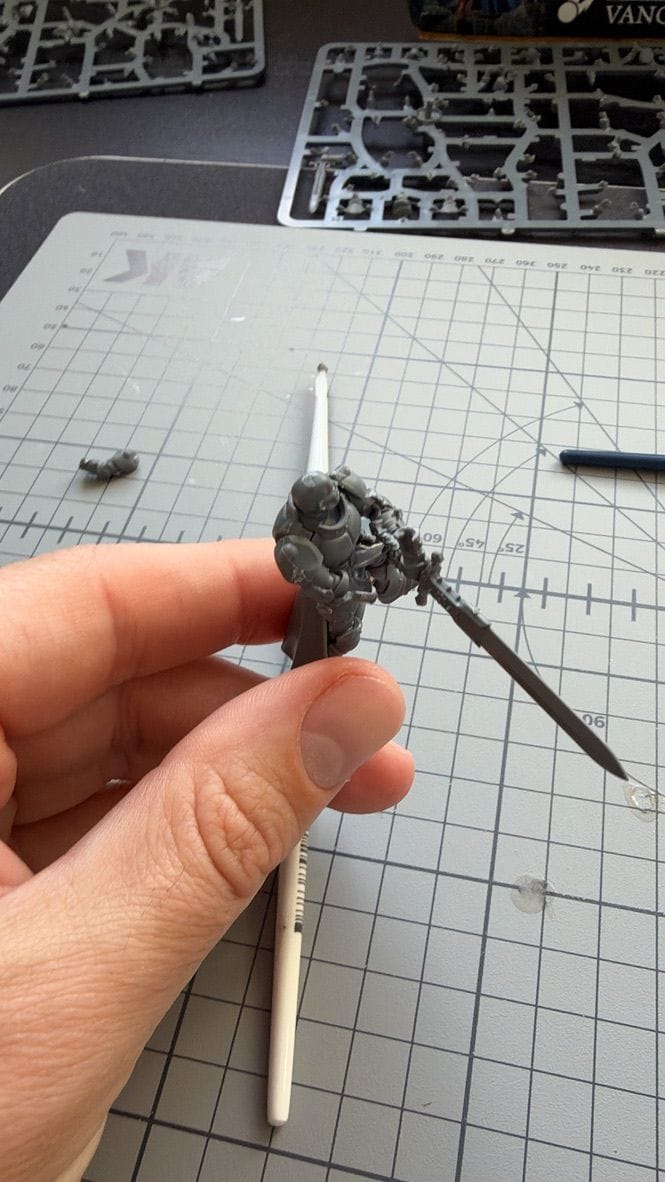 A built Vanquisher with a long sword, held up between finger and thumb over a cutting mat.